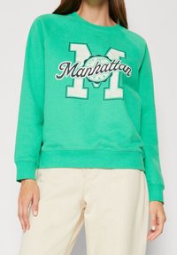 Mujer vistiendo un sudadera verde con la palabra "Manhattan" y una gran letra M, combinada con pantalones beige claros.