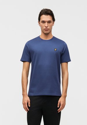 Muž v jednoduchom modrom tričku s krátkym rukávom a malým žltým logom vtáka na hrudi, stojaci pred jednoduchým svetlosivým pozadím.