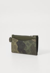 Green camouflage wallet with a textured finish, featuring a zippered top, a smooth leather accent, and a decorative metal ring.