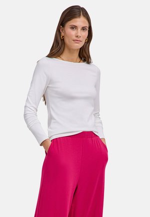 Frau trägt ein weißes Langarmshirt und knallpinkfarbene hoch taillierte Hosen, steht mit den Händen in den Taschen vor einem einfarbigen Hintergrund.