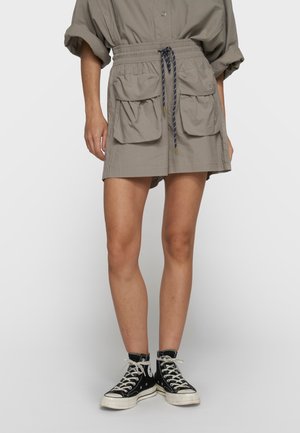 Femme portant une jupe taupe avec de grandes poches avant et une taille à cordon, associée à des baskets montantes noires avec des lacets blancs.