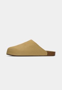 Beige slip-on klomper i semsket skinn med en glatt tekstur, rund tå, og en brun, strukturert såle. Minimalistisk design uten synlig maskinvare.