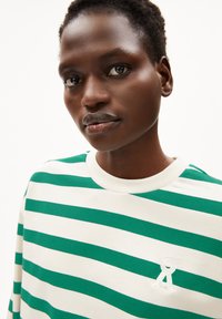 Nahaufnahme einer Person mit kurzem Haar, die ein weißes und grünes gestreiftes Hemd mit einem kleinen weißen Logo auf der Brust trägt.