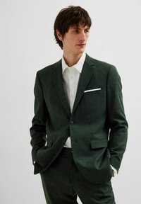 Tumši zaļš uzvalks ar teksturētu audumu, viena rindas pogas, divas pogas un iedobti atloki. Balta krekla ar plašu apkakli un krūšu kabatu.