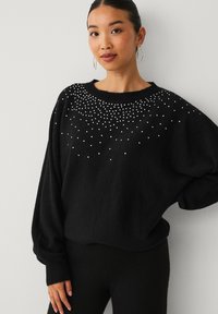 Sort oversized sweater med perleudsmykninger på den øverste front, ribbede manchetter og rund hals. Blød tekstur og løs pasform.