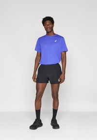 Ανδρική αθλητική ενδυμασία με μωβ κοντομάνικη μπλούζα και μαύρο σορτς. Το υλικό φαίνεται ελαφρύ. Μαύρες κάλτσες και αθλητικά παπούτσια ολοκληρώνουν την εμφάνιση.