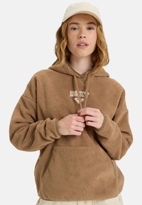 Ung kvinde, der justerer snoretrækkene på en brun fleece-hoodie med "Roxy"-logo, iført beige kasket og lyse bukser mod en hvid baggrund.