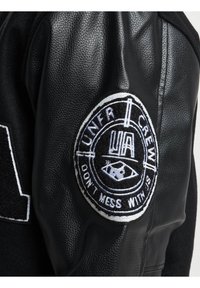 Veste en cuir noir avec un patch en laine sur l'épaule arborant un logo circulaire brodé. La texture est lisse avec une coupe ajustée.