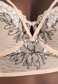 BH med gjennomsiktig nude-stoff og svart blonderbroderi, justerbare stropper, og en trekantet form med en sentral klaffdetalj.
