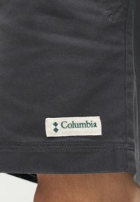 Σκούρο γκρι ύφασμα με ραμμένη ραφή και λευκή ορθογώνια ετικέτα της Columbia Sportswear Company ραμμένη κοντά στην κάτω άκρη.