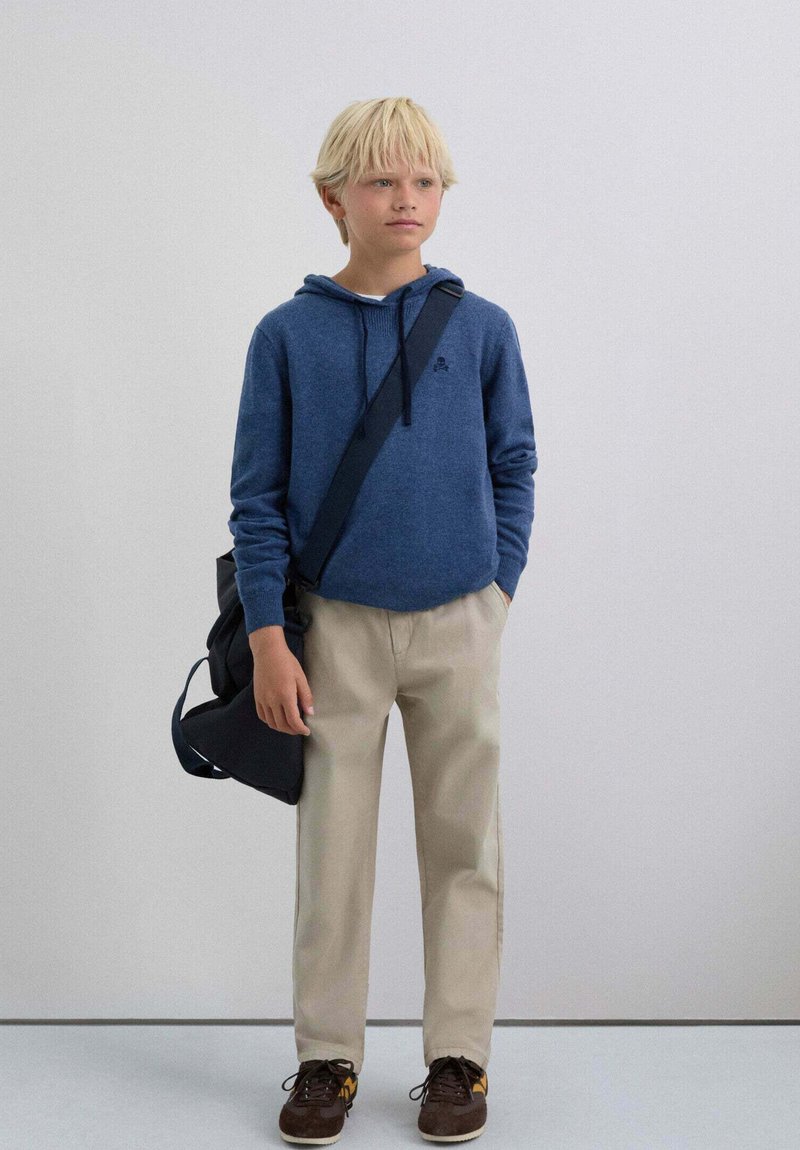 Sudadera con capucha de tela azul oscuro, pantalones beige, zapatos marrones y un bolso de hombro negro. Diseño sencillo con un bolsillo delantero y cordón ajustable.