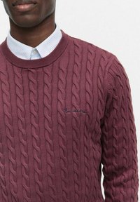 Vinröd kabelstickad tröja med rund hals och subtil brodyrdetalj. Bärs över en ljusblå skjorta med krage som visar upp ett strukturerat mönster.