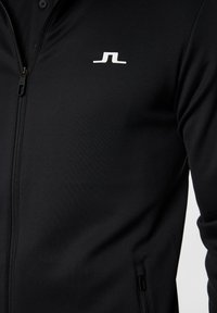 Svart zip-up jacka av mjukt, stretchigt material med en liten vit logotyp på bröstet och fickor med dragkedja på sidorna.