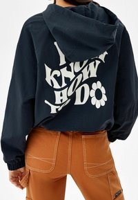 Osoba ubrana w czarną bluzę z kapturem z napisem "Wiesz jak to robisz" oraz graficznym motywem kwiatu z tyłu, połączona z brązowymi spodniami z widocznym ściegiem.