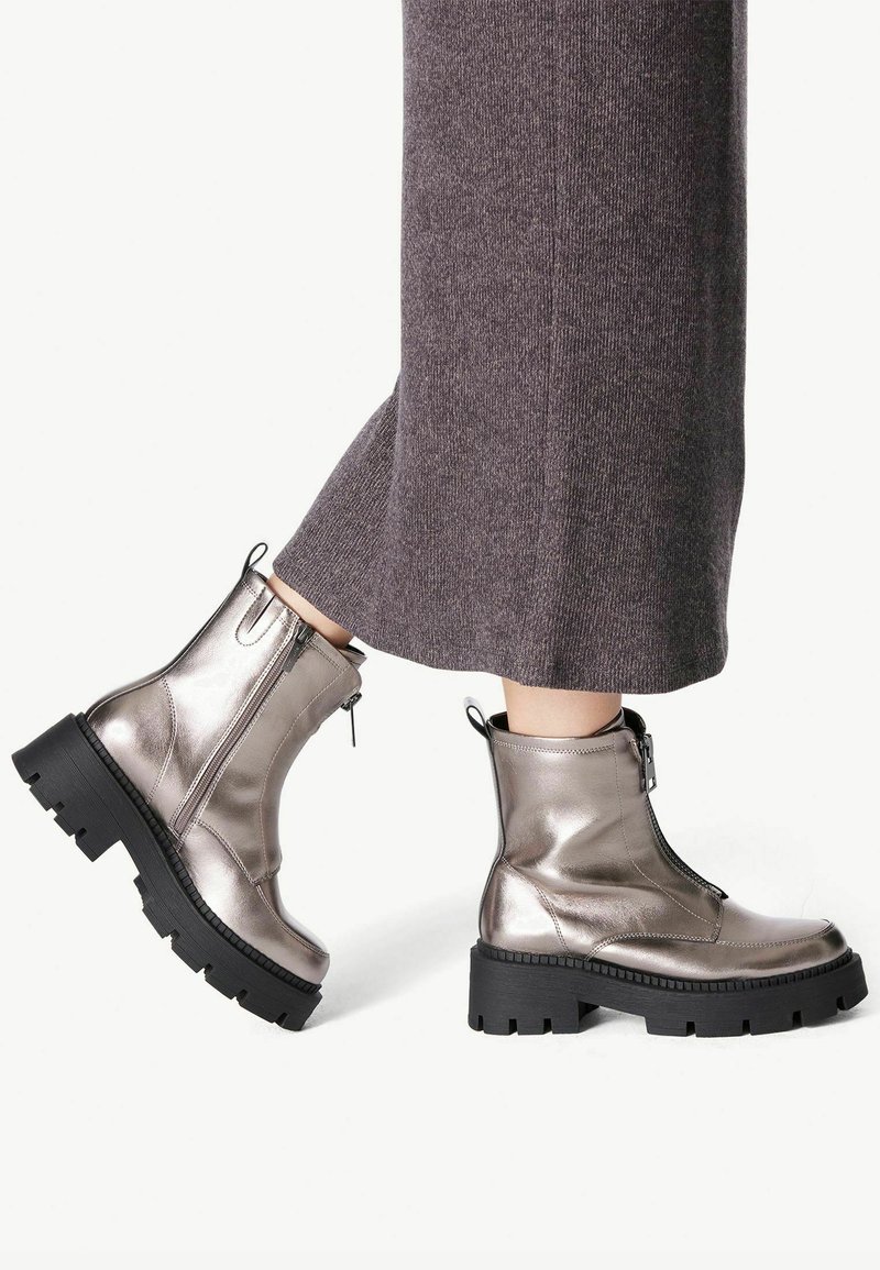 Botines metálicos plateados con una suela de goma negra gruesa, cierre lateral y detalle de tira para poner. Combinados con unos pantalones de punto grises.