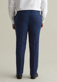 Pantaloni di lino blu con vestibilità sartoriale, dotati di due tasche posteriori e una texture liscia. Abbinati a scarpe eleganti nere, mostrati da dietro.