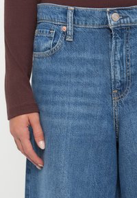 Jeans de mezclilla azul con cintura alta, cinco bolsillos y remaches de cobre. La tela tiene una textura suave y un ligero desgastado.