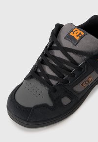Zapatilla de skate en negro y gris con parte superior de ante y malla, cierre de cordones, puntera redonda y logo de DC en naranja en la lengüeta.