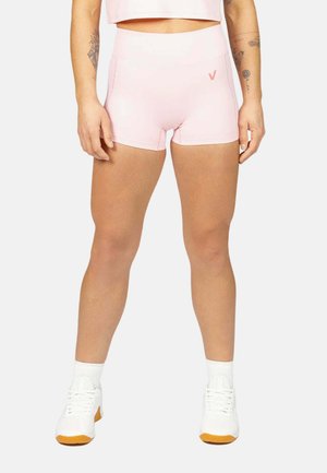 Persona con pantalones cortos ajustados rosa claro y top corto a juego, zapatillas blancas, de pie frente a un fondo liso.