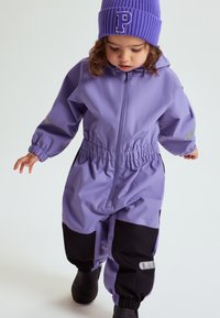 Peuter in een paarse en zwarte waterproof overall en een paarse muts met een "P", loopt tegen een effen achtergrond.