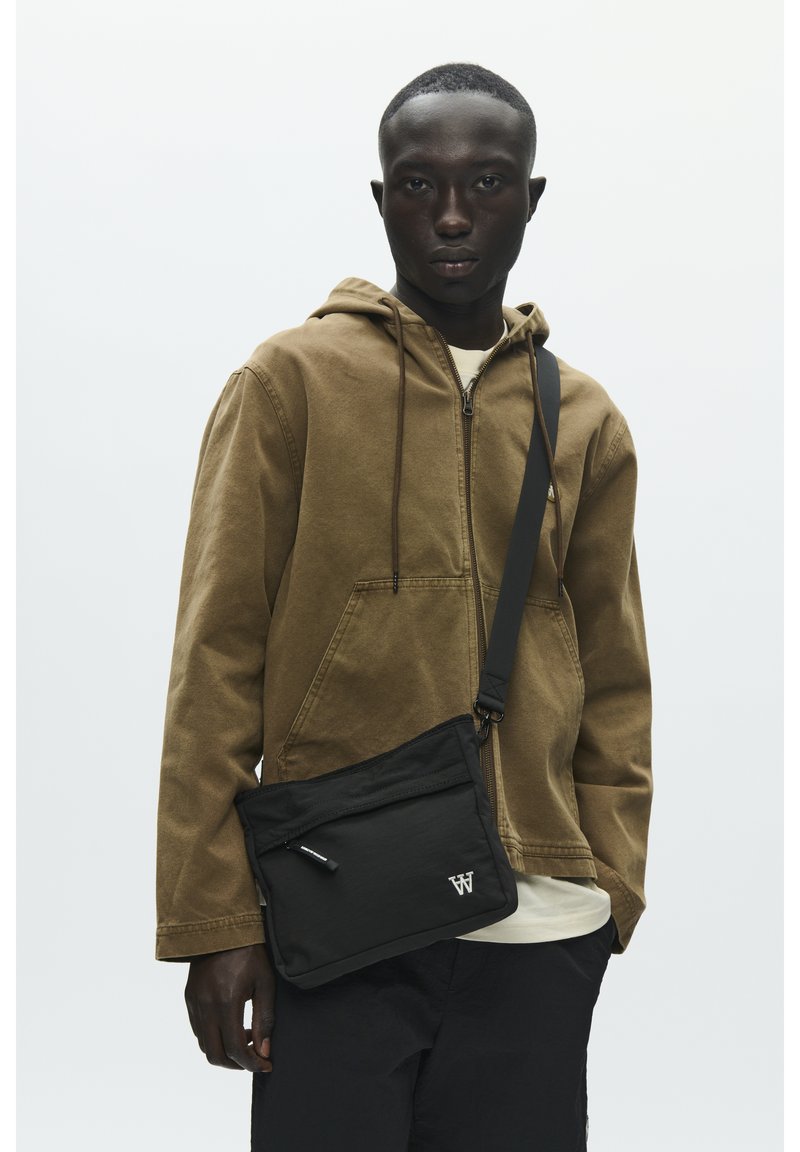 Sweat à capuche marron avec fermeture éclair et cordons, sac bandoulière noir avec une poche zippée, porté par-dessus une chemise blanche et un pantalon foncé.