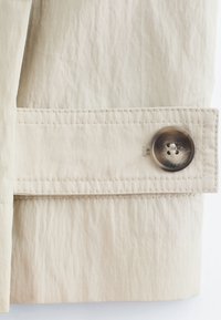 Passant de ceinture en tissu beige sur la manche d'un vêtement, fixé par un grand bouton marron à quatre trous avec des coutures détaillées.