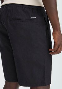 Shorts noirs avec une texture lisse, comportant une poche arrière et une étiquette de marque. La taille est élastique, offrant confort et flexibilité.