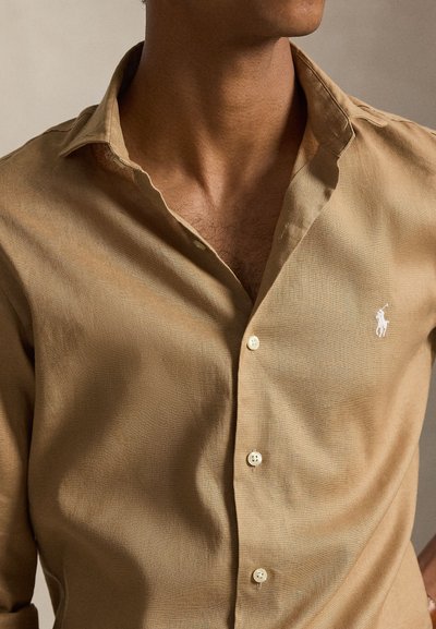 Άνδρας που φοράει μπεζ πουκάμισο με κουμπιά και ανοιχτό γιακά, με μικρό λευκό κεντητό λογότυπο παίκτη πόλο στο στήθος.