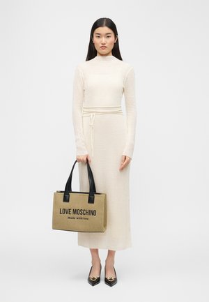 Une femme se tient debout tenant un sac cabas beige Love Moschino avec des poignées noires, portant une robe crème côtelée à manches longues et des chaussures pointues noires avec un détail doré.