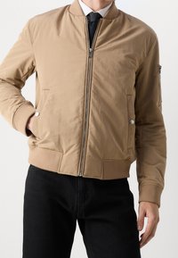 Tan bomberjacka med zipframslutning, ribbade cuffs och nederkant, sidofickor och en praktisk zipficka på vänster arm. Slät tygtextur.