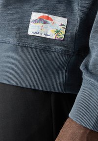 Blauwe geribbelde trui met een kleurrijke patch in strandthema, waarop palmbomen en een zon die ondergaat achter bergen zijn afgebeeld.