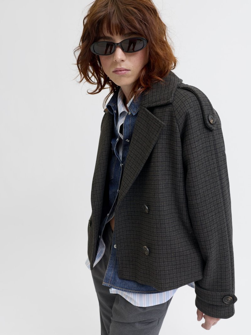 Chaqueta de cuadro oscuro de doble botonadura con grandes botones, tejido texturizado y un cuello. Usada en capas sobre una camisa de mezclilla y una camisa a rayas.