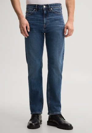 Mann trägt blaue gerade geschnittene Jeans und schwarze Lederschuhe und steht vor einem schlichten hellen Hintergrund.