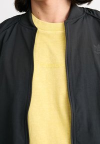 Svart bomberjacka med dragkedja, med en ribbad krage. Under den, en ljusgul sweatshirt i texturerad tyg med präglad logotyp.