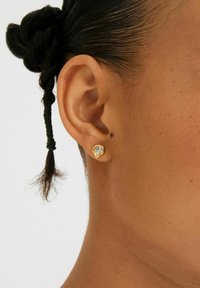 Nærbillede af en persons øre med en lille guld stud-ørering, der har en klar rund perle, og en kort fletning.