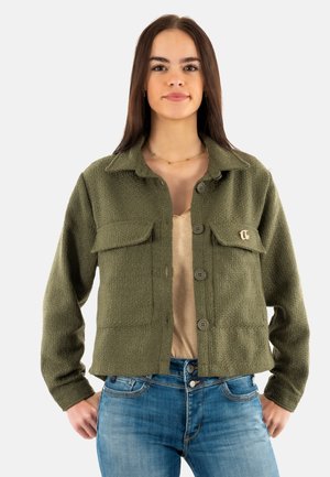 Veste texturée vert olive avec un col, fermetures à boutons à l'avant et deux poches poitrine, associée à un haut beige et un jean.