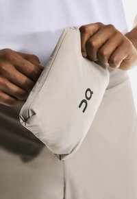 Des mains tenant un manteau plié de couleur beige clair avec le logo "On", sur un fond beige assorti.