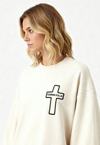 Femme aux cheveux blonds mi-longs portant un sweat-shirt blanc avec un patch noir en forme de croix portant l'inscription « KARMA KILLS », regardant vers la gauche sur un fond uni.