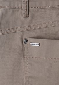 Gros plan sur la poche d’un pantalon kaki avec couture, passant de ceinture, rivet métallique et petite étiquette rectangulaire de la marque « Street One ».