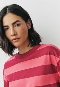 Junge Frau mit kurzem schwarzen Haar, die ein rot-rosa gestreiftes Shirt trägt und leicht zur Kamera schaut, vor einem schlichten Hintergrund.