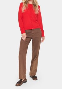 Cardigan rouge à manches longues avec des boutons et pantalon droit marron. Des chaussures en cuir marron avec des pampilles complètent la tenue.