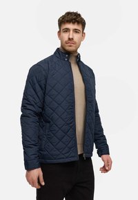 Marineblau gesteppte Jacke mit hohem Kragen, Reißverschluss vorne und seitlichen Taschen, kombiniert mit einem beigen Pullover darunter.