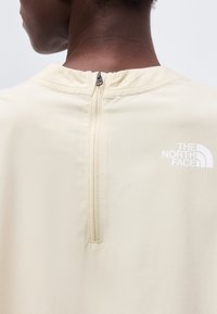 Πλάγια όψη ατόμου που φοράει τοπ μπεζ με φερμουάρ στο πίσω μέρος και λευκό λογότυπο The North Face στο επάνω δεξιό μέρος.