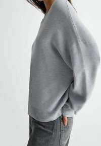 Šedý oversized svetr s širokým výstřihem a žebrovaným lemem. Materiál má hladkou strukturu, doplněnou jemnými detaily.