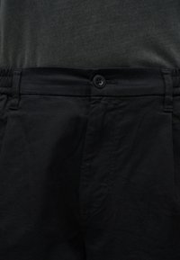 Μαύρο παντελόνι στην περιοχή της μέσης με κλείσιμο με κουμπιά και θηλιές για ζώνη, φορεμένο με σκούρο γκρι πουκάμισο μέσα μέσα.