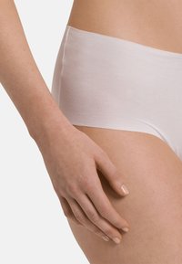 Sottovesti hipster bianche in cotone con bordo liscio e senza cuciture. La mano del modello è appoggiata delicatamente sulla coscia, mettendo in mostra un tono di pelle naturale.