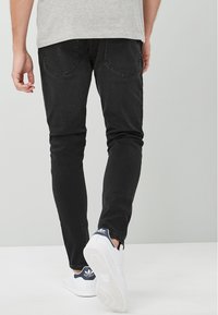 Vaqueros negros de corte slim con diseño de cinco bolsillos, detalles de costura y una textura suave, combinados con zapatillas blancas y una camiseta gris.