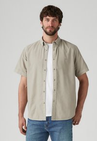 Light beige short-sleeve shirt with button front, collar, and subtle logo, paired with a white t-shirt and blue jeans.