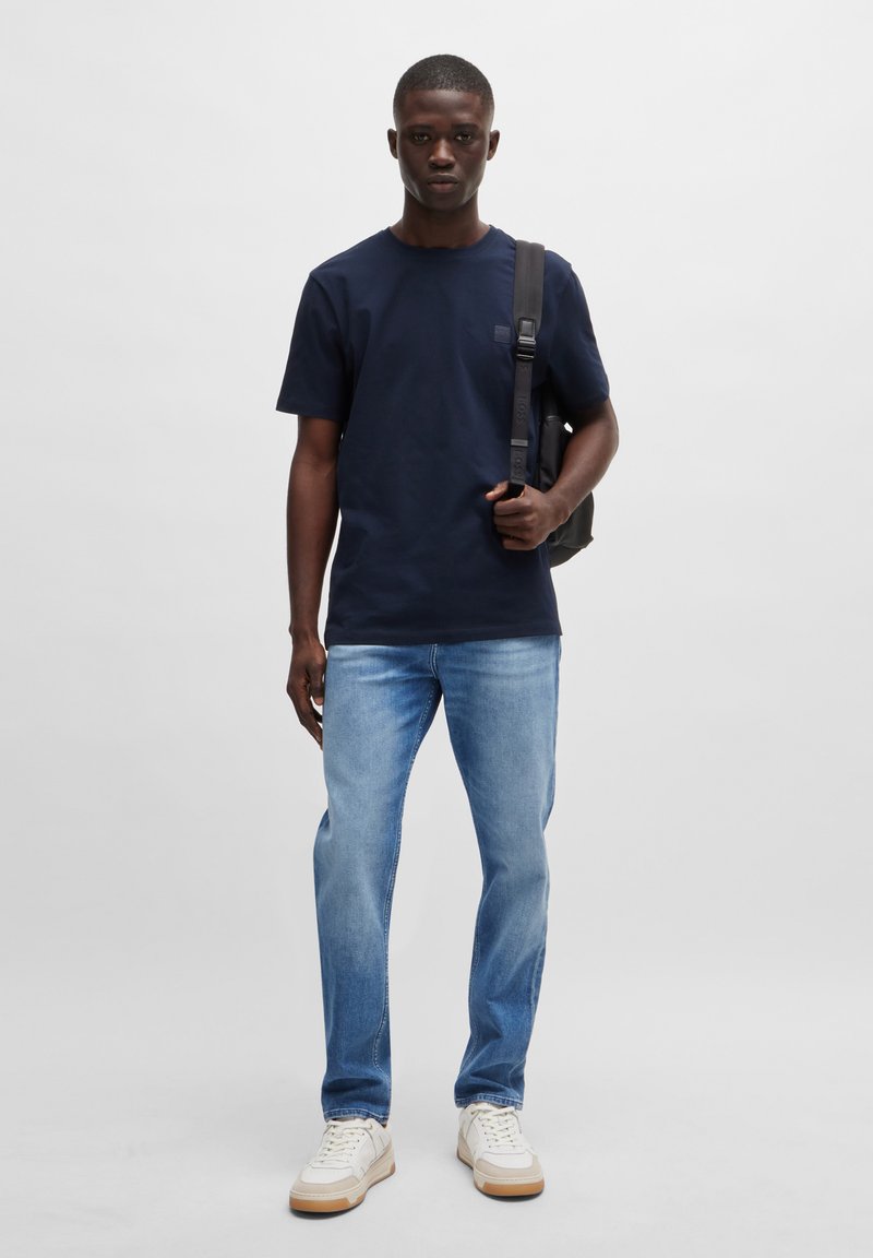 T-shirt en coton bleu marine, jean droit bleu clair, baskets blanches avec semelles en caoutchouc, et un sac à dos noir porté sur une épaule.