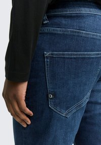 Blugi bleu închis cu un design ajustat, având un buzunar pe spate și un mic eticheta neagră. Materialul pare neted, cu un ușor luciu.